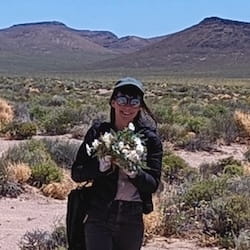 A smiling person stands in a sunny landscape.